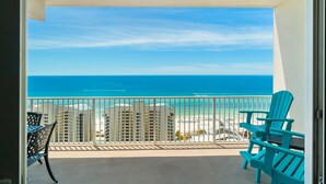 A balcony with a view of the ocean and beach, featuring two Adirondack chairs and a table.