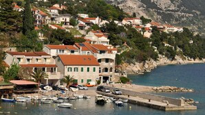 Un pueblo costero con edificios a lo largo del paseo marítimo, un puerto deportivo con barcos y una ladera rocosa al fondo.
