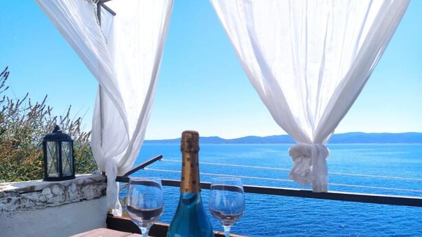 Una mesa de madera con una botella de vino azul y dos copas, frente a un mar azul cristalino y montañas distantes.