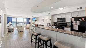 A modern kitchen with white cabinetry, a granite countertop, and a black refrigerator. There are two bar stools and a dining area with a view of the ocean. - The Oceans 405- 2 Bed OF Condo with Pool (North Myrtle Beach)