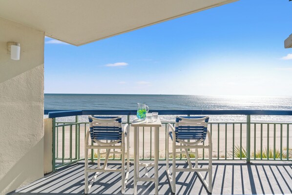 A balcony with two chairs and a table, overlooking a beach and the ocean.