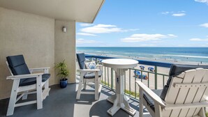 A balcony with two white chairs, a small table, and a potted plant. The balcony overlooks a beach with people and umbrellas.
