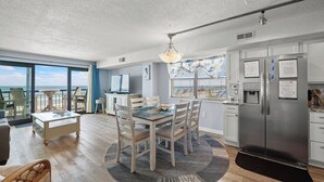 A modern kitchen with a large refrigerator, dining table, and chairs. There is a view of the ocean through large windows.