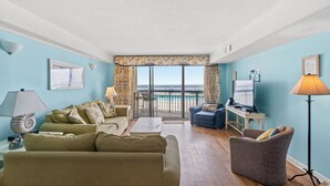 Un salon avec un canapé, un fauteuil et une table sur laquelle se trouve un ordinateur portable. Une grande fenêtre offre une vue sur la mer.