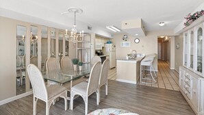 A spacious kitchen with a central island, a dining area with a glass table and wicker chairs, and a living area with a sofa and a TV.