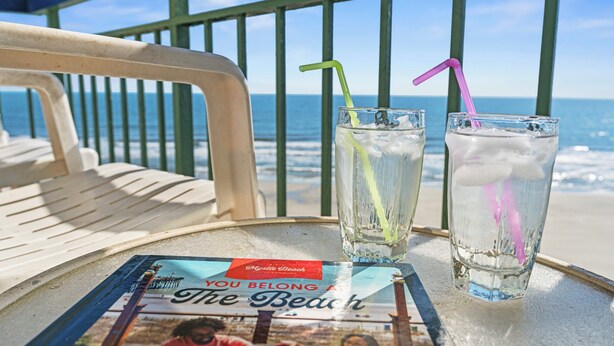 Two glasses of iced drinks with straws on a table with a magazine titled "You Belong to the Beach" and a chair facing the ocean.