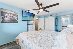 Ein Schlafzimmer mit einem Bett, Deckenventilator, Fernseher, Kommode, Pflanze und einer Tür.