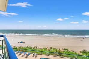 Blick auf einen Strand vom Balkon aus, mit Liegestühlen, einem Rettungsschwimmer-Schild und einem Pool.