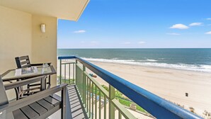 A balcony with a view of the beach, a table with chairs, and a railing.