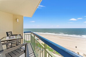 A balcony with a view of the beach, a table with chairs, and a railing.