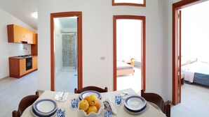 Een eethoek met een tafel gedekt voor twee personen, een keuken met houten keukenkastjes en zicht op een slaapkamer door een deuropening.