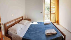 Een eenpersoonsbed met houten hoofdbord, witte beddengoed en een blauwe sprei. Er is een raam met houten luiken en uitzicht op groen.