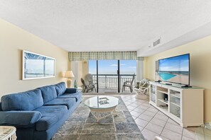 Una sala de estar con un sofá azul, una mesa de centro de vidrio, un televisor sobre un mueble bajo y un balcón con vista al mar.
