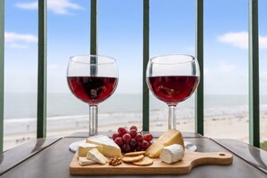 Twee glazen rode wijn, een selectie kazen, druiven en crackers op een houten plank, met op de achtergrond een uitzicht op het strand en de zee.