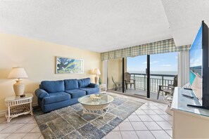 A living room with a blue sofa, a glass coffee table, and a view of the ocean through a balcony. - Oceans 814 - Ocean Front 1 Bedroom Condo in Cherry Grove (North Myrtle Beach)