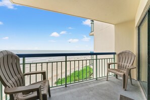 A balcony with two chairs, a view of the beach, and a clear sky.