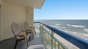 Een balkon met twee stoelen en een tafel, met uitzicht op een strand met mensen en boten.