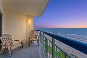 Een balkon met twee stoelen en een tafeltje, met uitzicht op een strand en een heldere horizon van de oceaan.