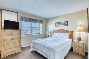A bedroom with a bed, a dresser, a television, a window with blinds, a nightstand with a lamp, and a wall decoration.