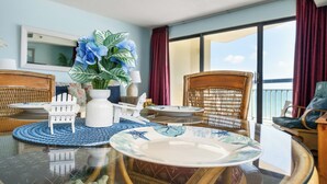 A dining area with a glass table, wicker chairs, a white decorative chair, and a view of the sea through large windows. - Oceans 816 - Great Views - 1 Bedroom Ocean Front Condo (North Myrtle Beach)