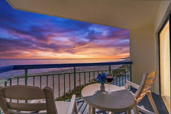 A balcony with a table and chairs, overlooking a beach with a sunset view.