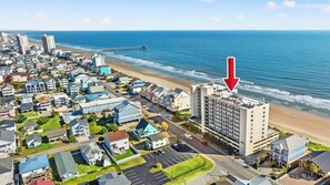 An aerial view of a coastal town with a prominent hotel near the beach, featuring a parking lot and a view of the ocean.