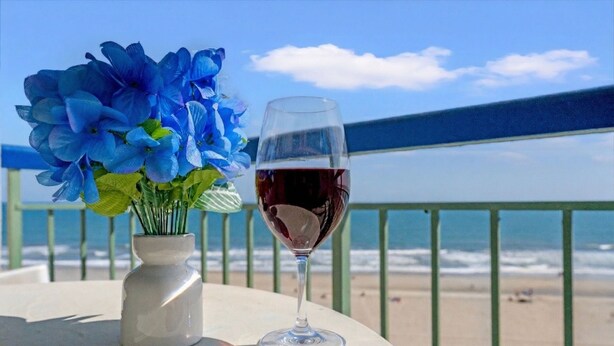 A table with a vase of flowers and a glass of red wine, overlooking a beach with a railing. - Oceans 816 - Great Views - 1 Bedroom Ocean Front Condo (North Myrtle Beach)