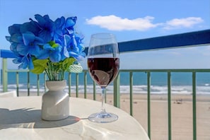 A table with a vase of flowers and a glass of red wine, overlooking a beach with a railing.