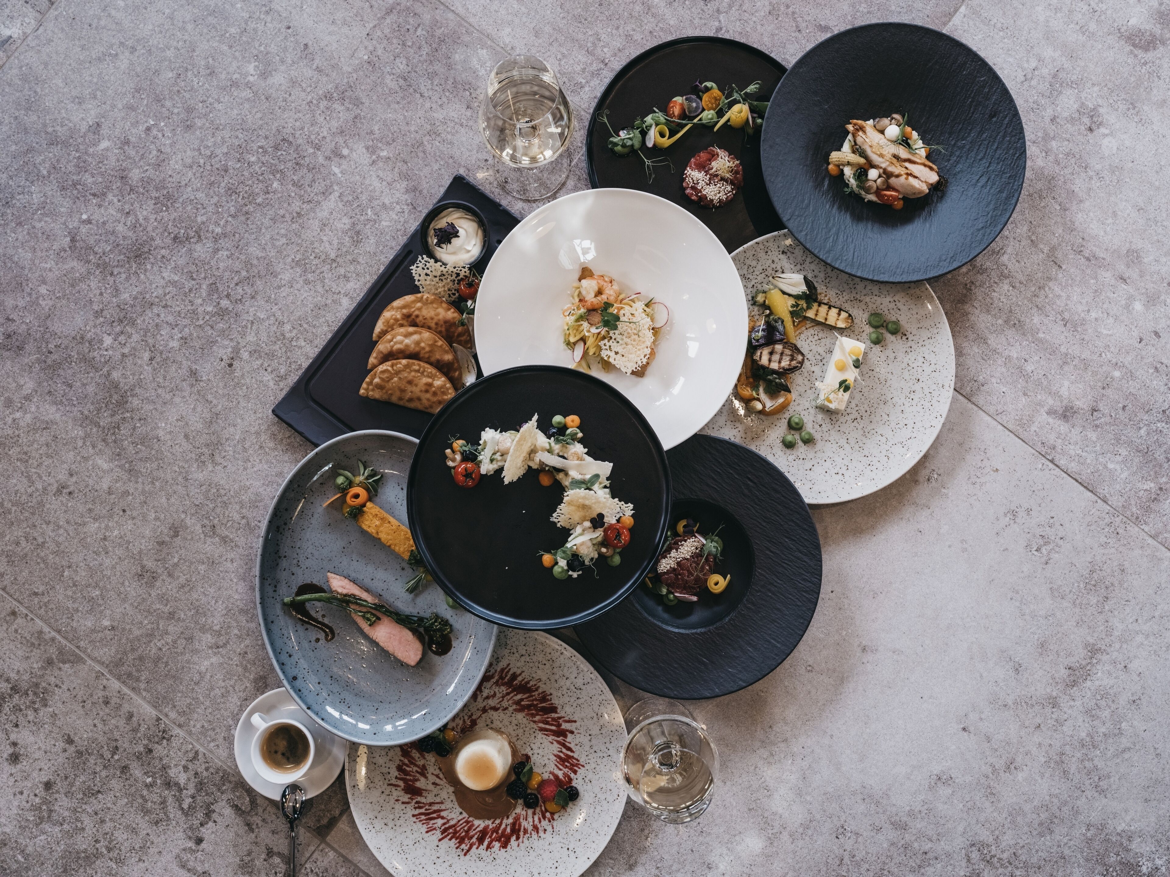 A top-down view of a dining table with various plates of food, glasses of water, and a cup of coffee.