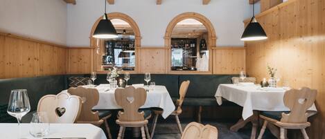 Interior de restaurante com paredes de madeira, janelas em arco e cadeiras em formato de coração. Mesas dispostas com toalhas brancas e taças de vinho.