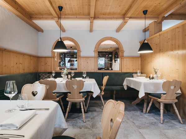 Interior de restaurante com paredes de madeira, janelas em arco e cadeiras em formato de coração. Mesas dispostas com toalhas brancas e taças de vinho.