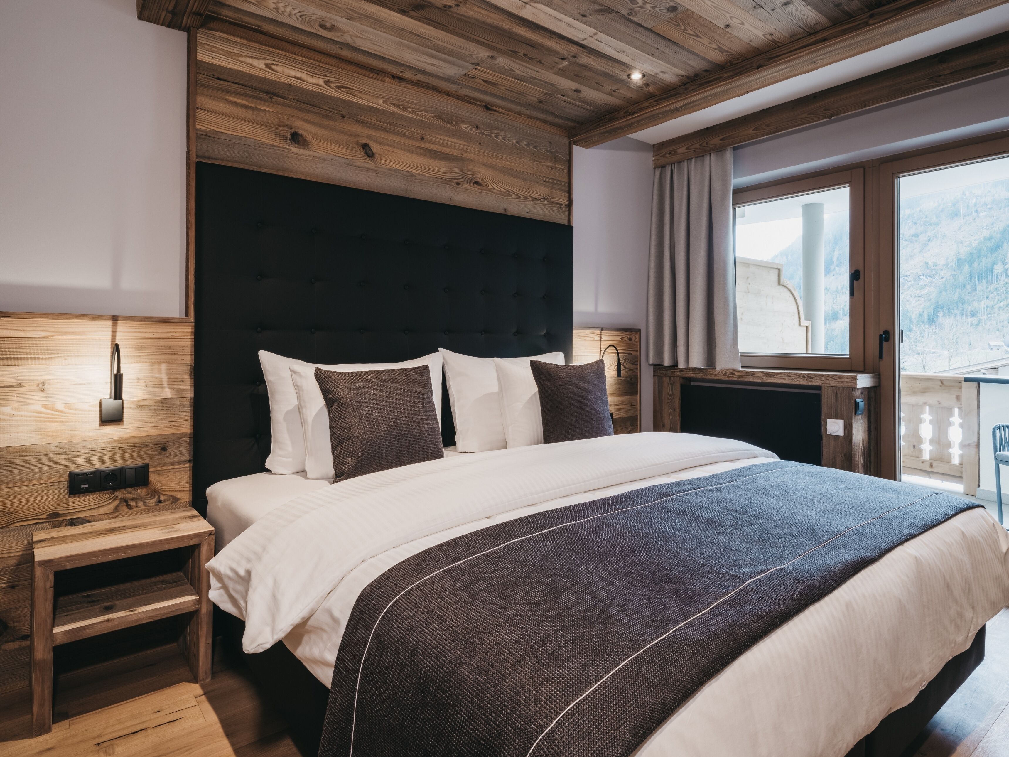 A bedroom with a wooden ceiling, a large bed with white and dark pillows, a nightstand, and a window with a view of a snowy landscape.