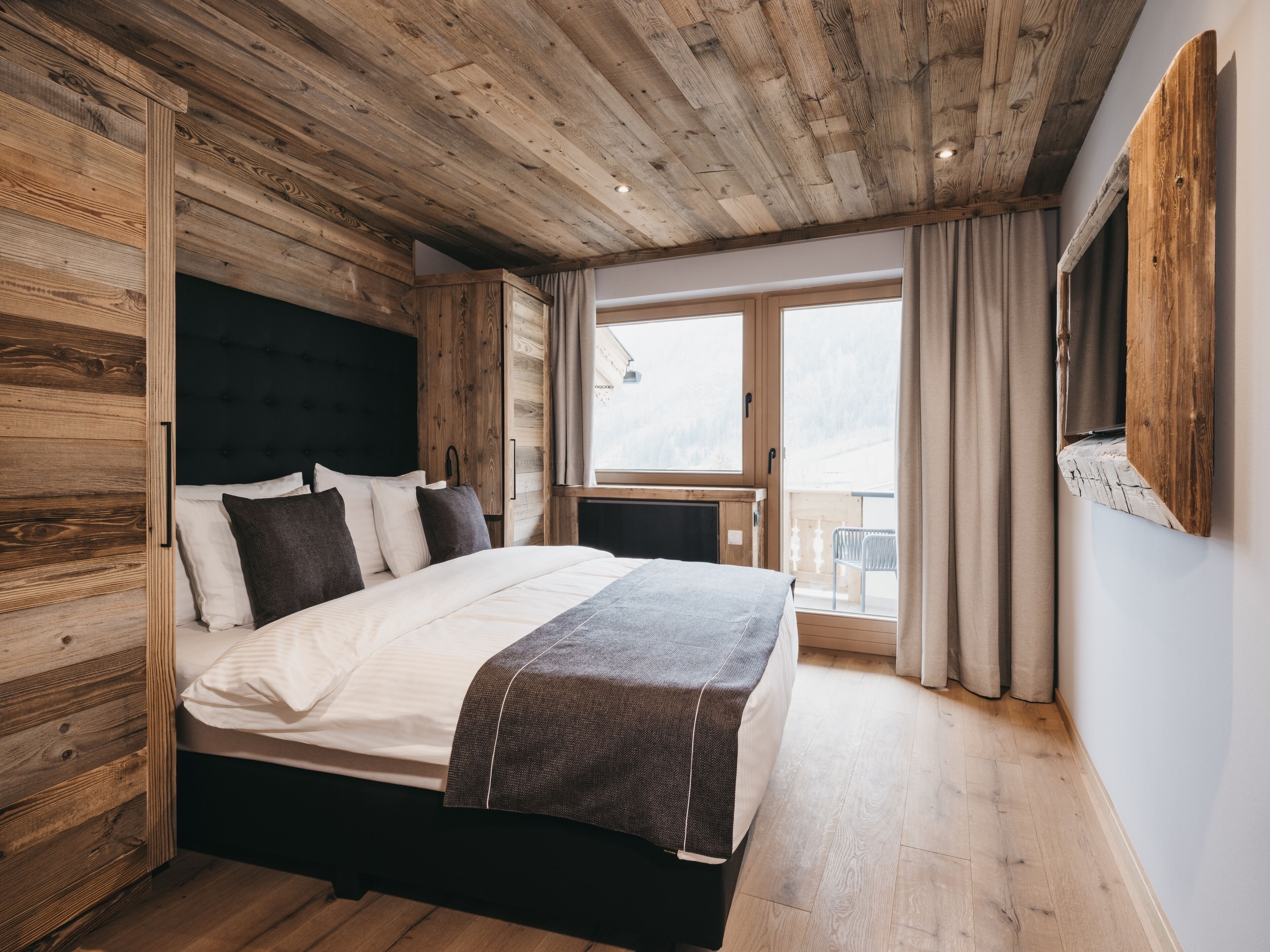 A bedroom with a wooden ceiling, a bed with a dark headboard, a window with curtains, and a view of the outdoors.