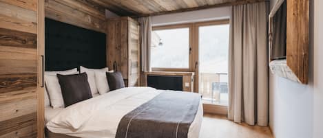 A bedroom with a wooden ceiling, a bed with a dark headboard, a window with curtains, and a view of the outdoors.