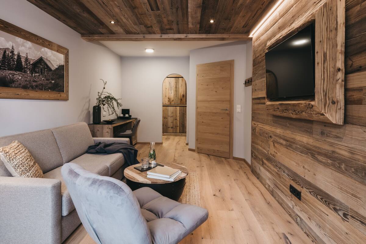 Een gezellige woonkamer met een houten plafond, een grote tv aan de muur, een bank, een salontafel en een houten deur.