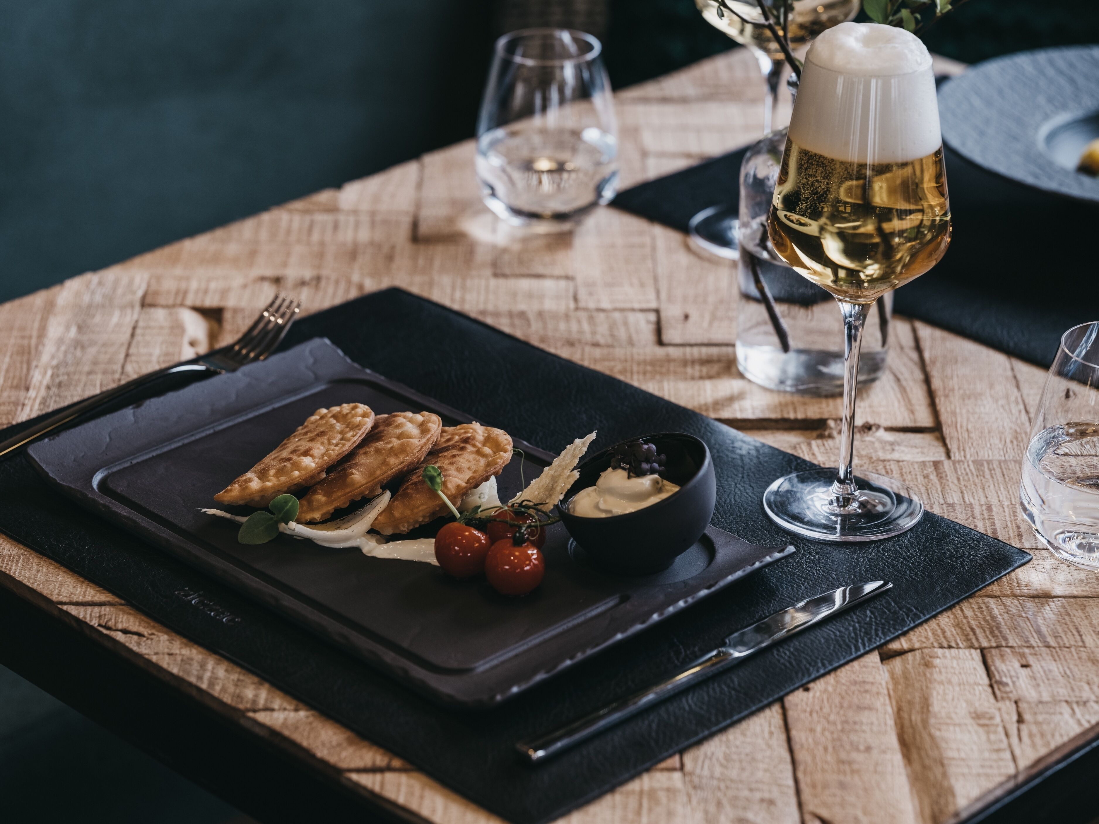 A dining table with a black placemat, a plate of food, a bowl of sauce, and a glass of white wine.