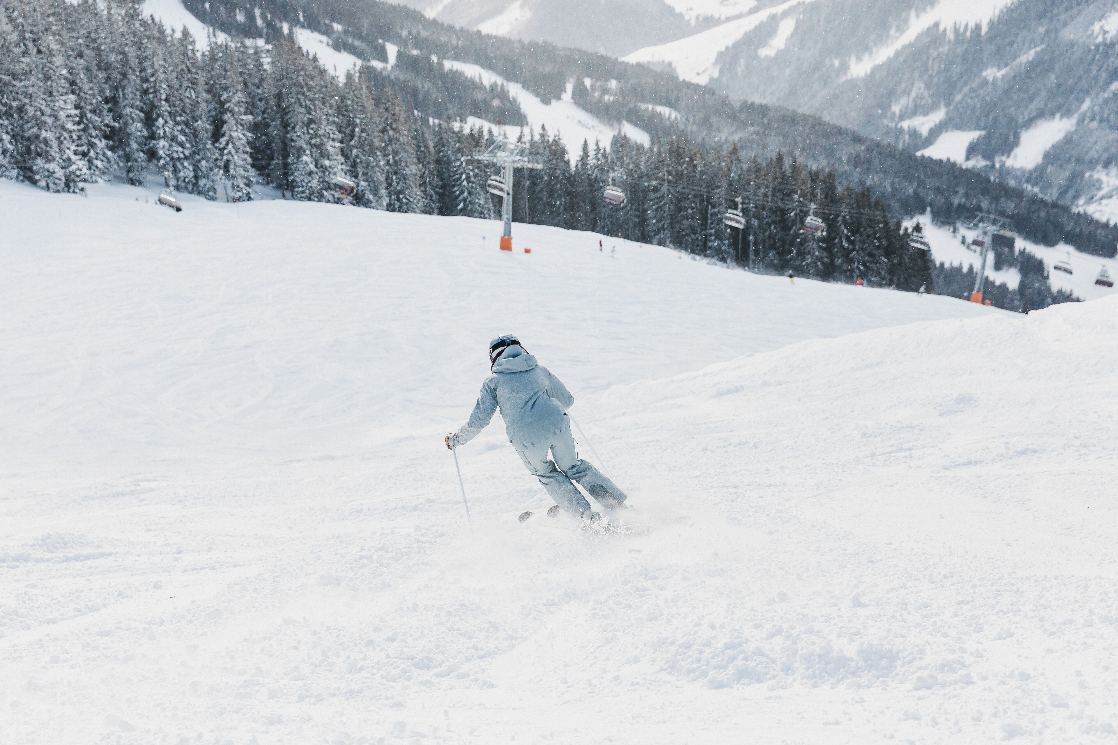 Une personne fait du ski sur une pente enneigée, avec des arbres et des montagnes en arrière-plan.