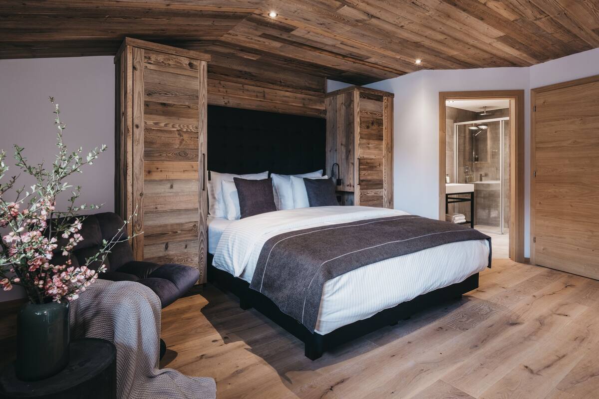 Een slaapkamer met een houten plafond, een groot bed met hoofdeinde, een nachtkastje, een vaas met bloemen en een zichtbare badkamer door een geopende deur.