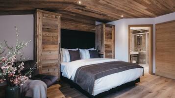 Un dormitorio con techo de madera, una cama grande con cabecera, una mesita de noche, un jarrón con flores y un baño visible a través de una puerta abierta.