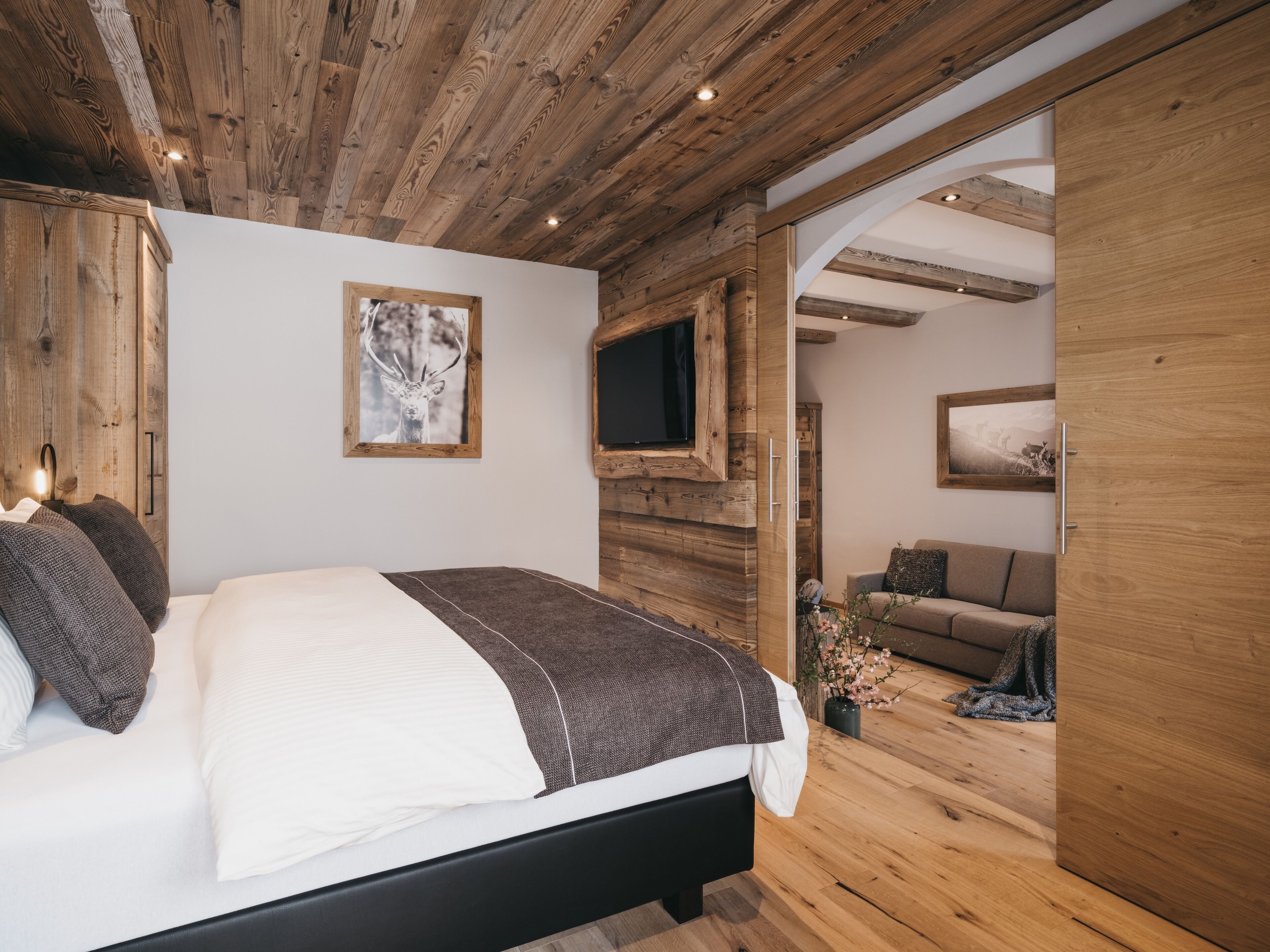 Un dormitorio con techo de madera, una cama con ropa de cama blanca y marrón, un televisor montado y un armario de madera.