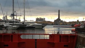 Un port de plaisance où sont amarrés plusieurs yachts, un phare imposant en arrière-plan et une barrière rouge au premier plan.