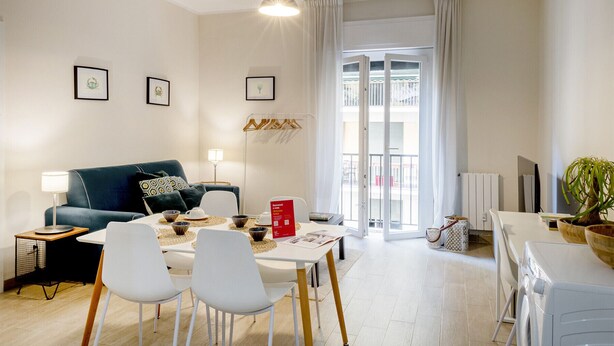 Een moderne woonkamer met een blauwe bank, witte stoelen, een eettafel en een groot raam met gordijnen.