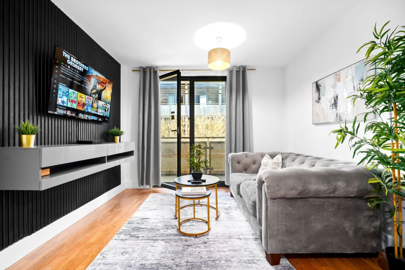 Un salon moderne avec un canapé gris, un téléviseur fixé sur un mur noir, une petite table ronde avec un cadre doré et une plante en pot.