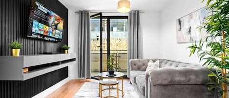 En moderne stue med en grå sofa, et tv monteret på en sort væg, et lille rundt bord med gylden ramme og en potteplante.
