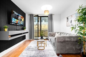 A modern living room with a grey sofa, a TV mounted on a black wall, a small round table with a gold frame, and a potted plant. - Arcadian Apartment - Secure Parking - Balcony (West Midlands)
