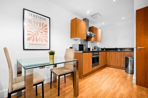 A modern kitchen with wooden cabinets, a glass dining table, and a framed artwork on the wall.