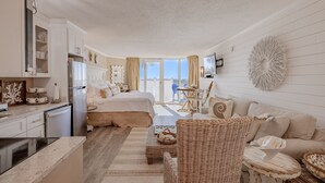 Een ruime slaapkamer met een groot bed, een zithoek met een bank en stoelen, een eettafel en een keuken met een koelkast en kastjes.