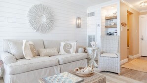 Een woonkamer met een beige bank, een ronde wanddecoratie, een klein tafeltje met een mand en een rieten stoel.