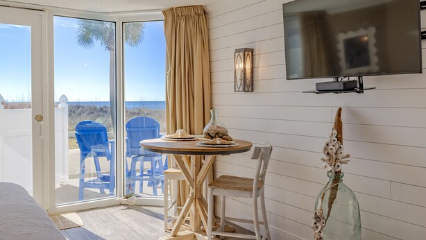 Een kamer met een schuifglazen deur naar uitzicht op een strand, een wandtelevisie, een kleine ronde tafel met stoelen en een decoratieve fles op de vloer.