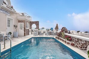 A pool area with a stone wall, a white building with a sign, and outdoor furniture. - Merovigliosso Budget Studio ground floor (Thira - Santorini)